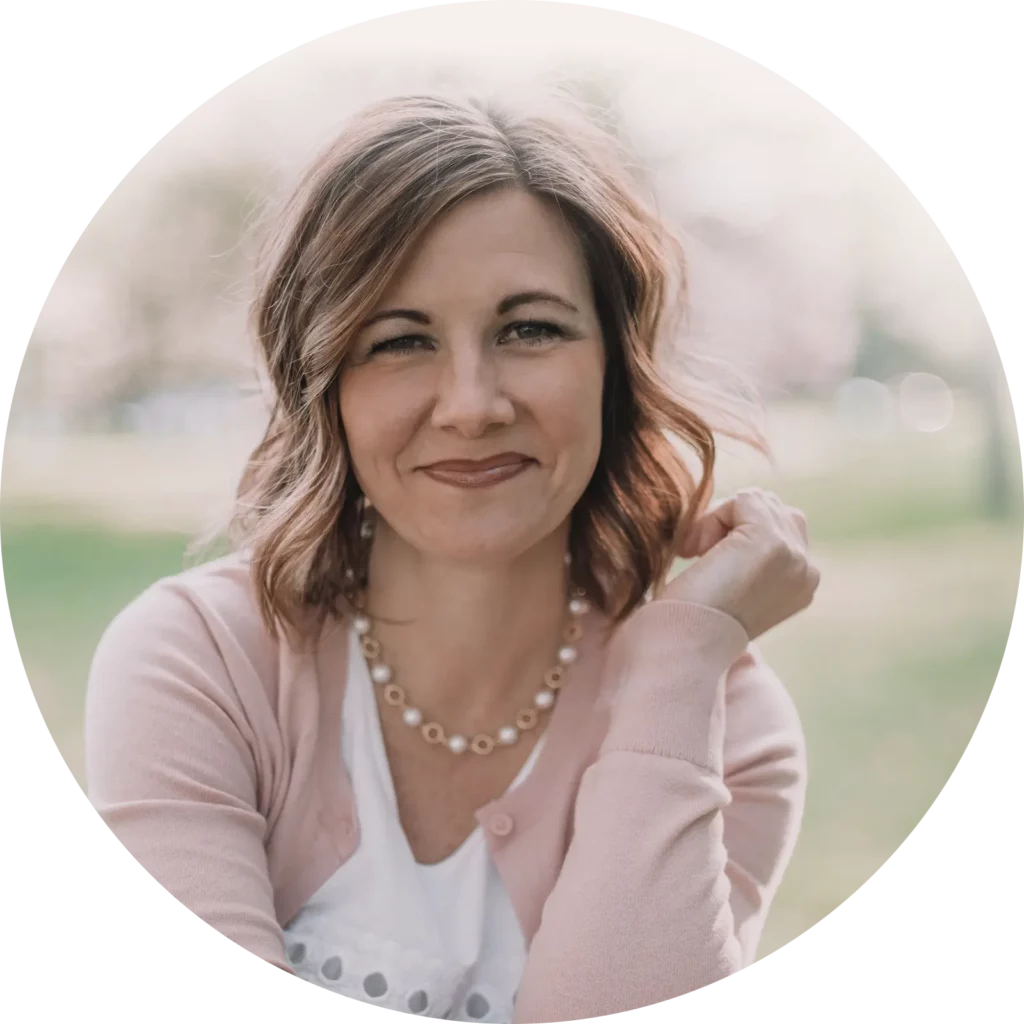 Molly smiling gently while seated outdoors, wearing a light pink cardigan and pearl necklace, with soft natural lighting and a blurred background of grass and trees.