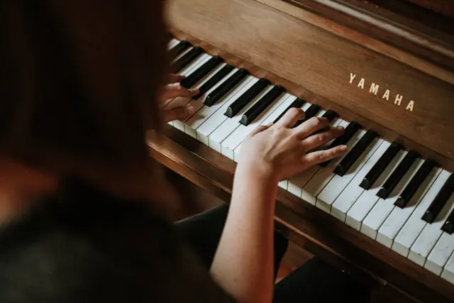 Person playing a Yamaha upright piano, with hands positioned on the keys and warm natural lighting highlighting the wooden instrument.